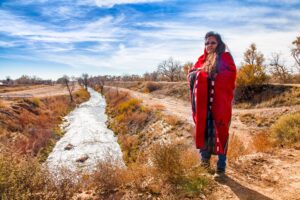 Native American woman wrapped in traditional blanket standing by acequia.